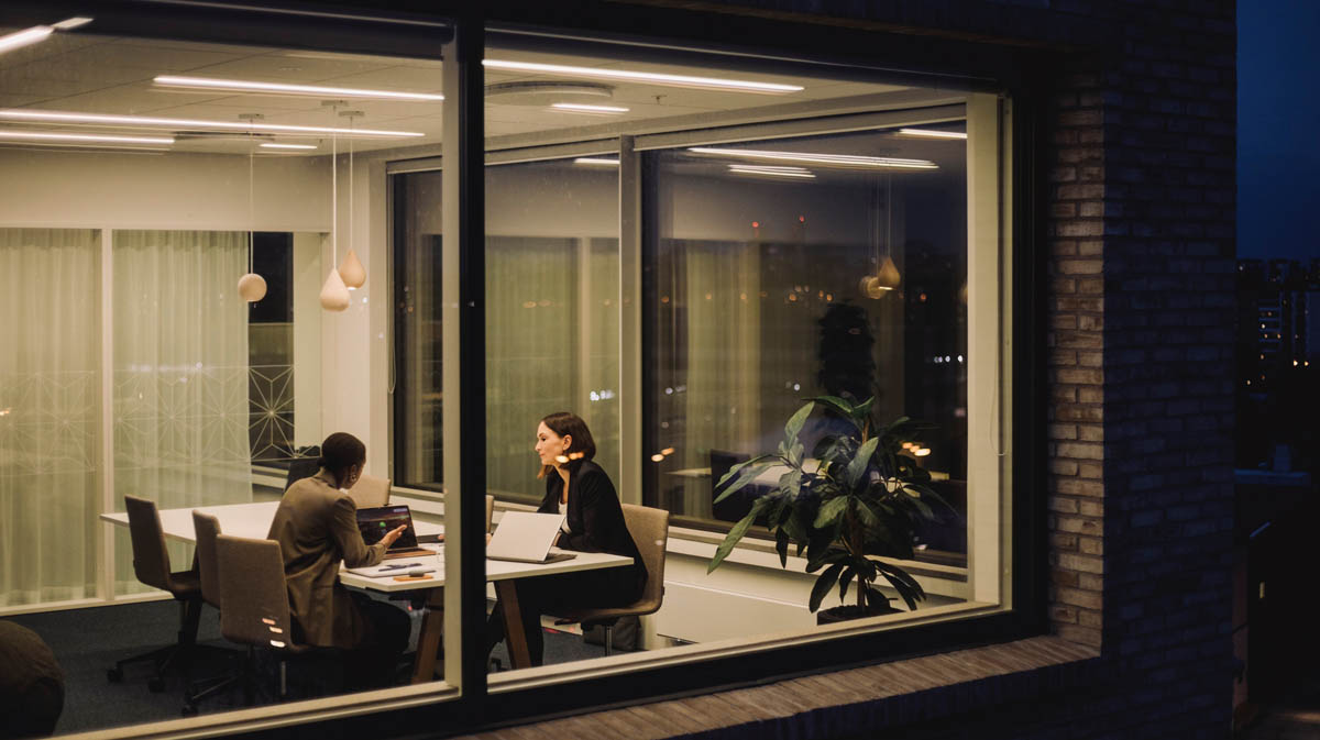 Två personer sitter i ett mötesrum på kontor och arbetar med laptop, sett genom fönster på kvällen
